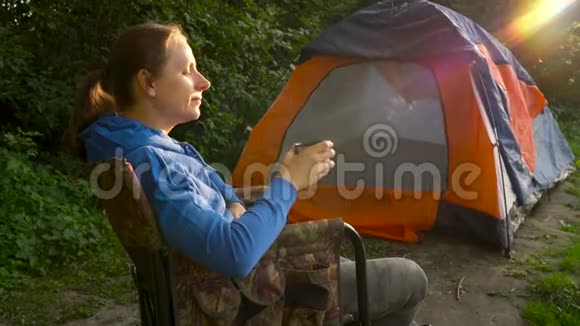 女人正在河边一个帐篷附近的扶手椅上喝茶视频的预览图