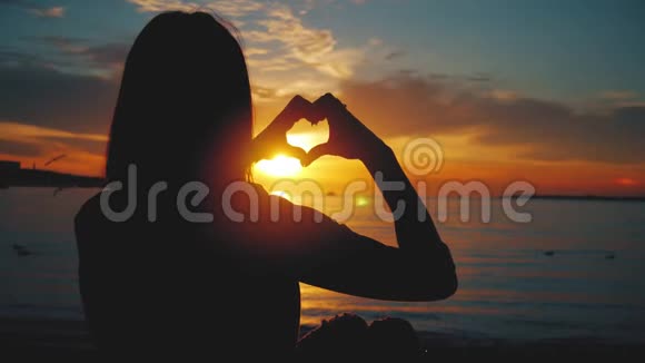 年轻美丽的女孩用她的双手在心形框架下夕阳在海洋上空情感观念视频的预览图