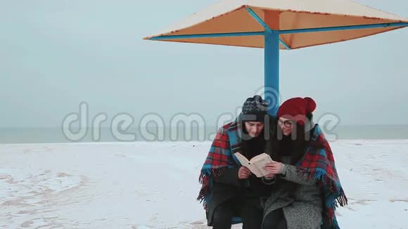 一个女孩和一个男人坐在海滩的天篷下盖着毯子看书视频的预览图