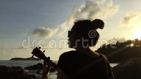 年轻的混血女孩在海滩上玩四弦琴五彩缤纷的日落高清后景银色泰国视频的预览图