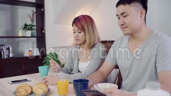 漂亮的亚洲年轻夫妇在吃早餐时一边拿着报纸和手机一边在桌子上分心视频的预览图