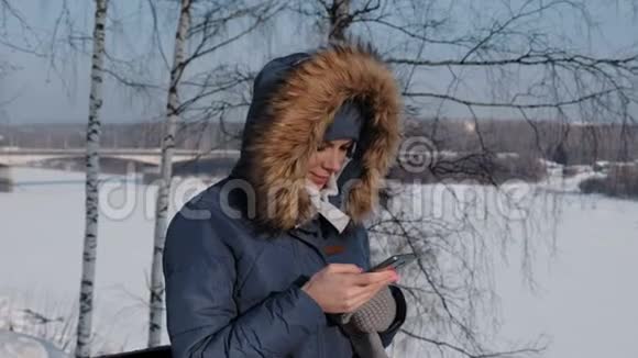 穿着蓝色羽绒服带着毛皮罩的女人在冬天公园的手机里写短信视频的预览图