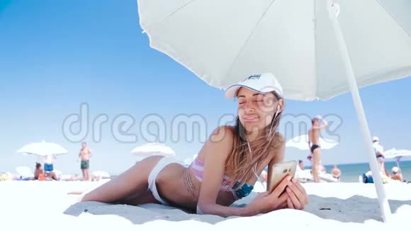 美丽的晒黑微笑的女人躺在沙滩上躺在沙滩上一边放松一边听着电话里的音乐视频的预览图