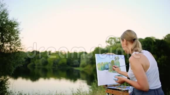 年轻艺术家在公园里用画笔和色彩坐在浪漫的湖泊和城市建筑旁视频的预览图