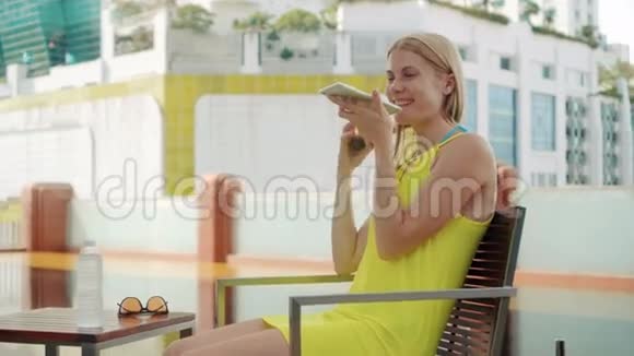 穿黄色衣服的女人坐在游泳池附近的椅子上用她的智能手机和朋友聊天视频的预览图