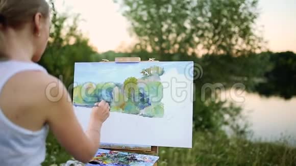 年轻艺术家在公园里用画笔和色彩坐在浪漫的湖泊和城市建筑旁视频的预览图