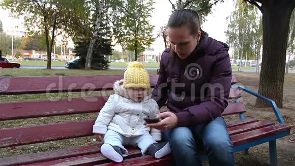 妈妈带着孩子坐在公园的长凳上妈妈给你的孩子看一部智能手机视频的预览图