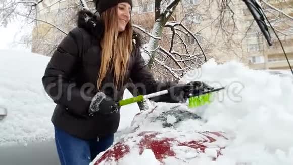 4k视频美丽微笑的年轻女子用刷子从雪地上清洁汽车挡风玻璃视频的预览图