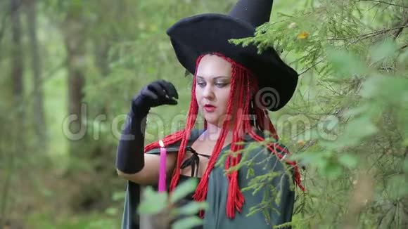 带着红色头发尖尖帽子和黑色斗篷的迷人的年轻女巫在森林里点燃蜡烛视频的预览图