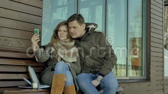 在一个坐在长凳上的春天公园里年轻的女人和男人在电话里自拍喝着热水瓶里的茶视频的预览图
