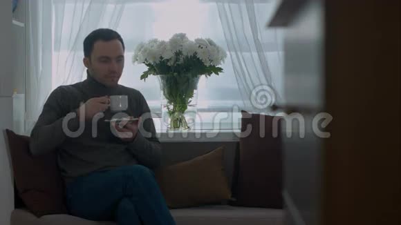 男人在沙发上喝咖啡在家透过窗户看着视频的预览图