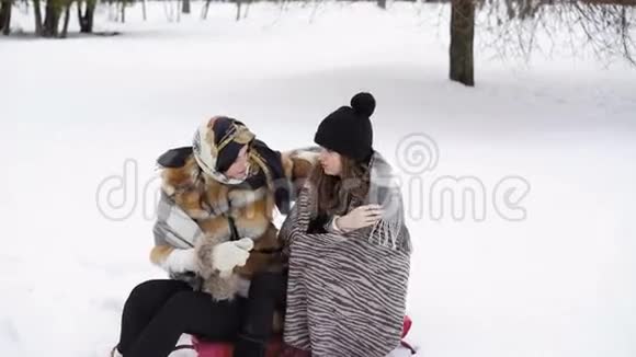 两个女孩穿着温暖的格子布坐在冬季公园4K视频的预览图