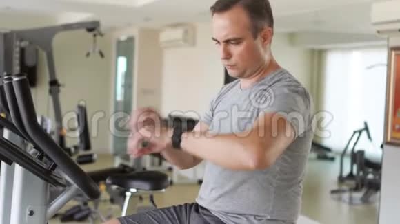一个英俊健康的年轻人在健身房骑着健身车做运动用他的智能手表检查生命体征视频的预览图