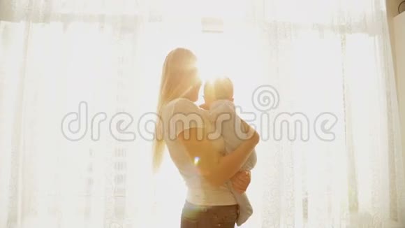美丽的年轻母亲在灿烂的阳光下抱着她的孩子在大窗户前视频的预览图