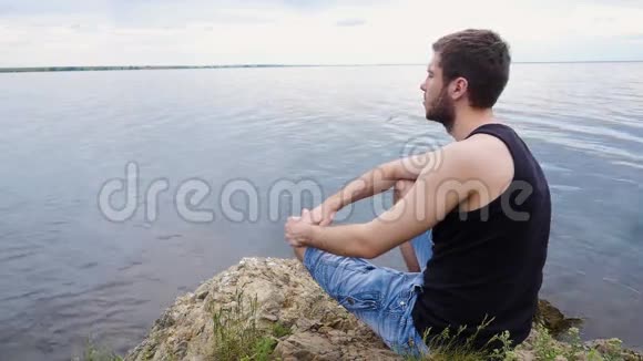 年轻人坐在一个乡村湖泊上方的岩石悬崖上乡村男孩孤独的年轻人视频的预览图