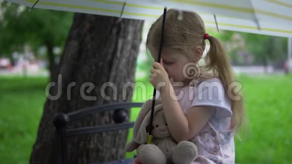 一个带着玩具兔子的小女孩坐在伞下的长凳上视频的预览图
