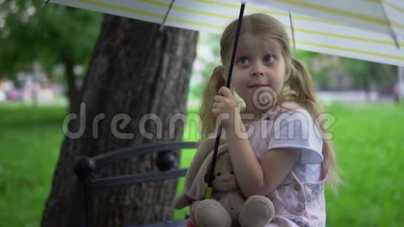 一个带着玩具兔子的小女孩坐在伞下的长凳上视频的预览图