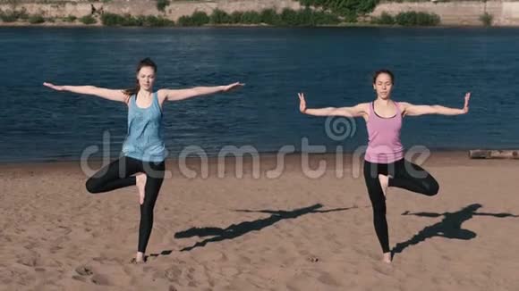两个女人站在城市河边的海滩上伸展瑜伽美丽的城市景色平衡姿势视频的预览图