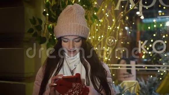 美丽迷人的女孩站在一家装饰有花环的橱窗附近使用智能手机新年圣诞节视频的预览图