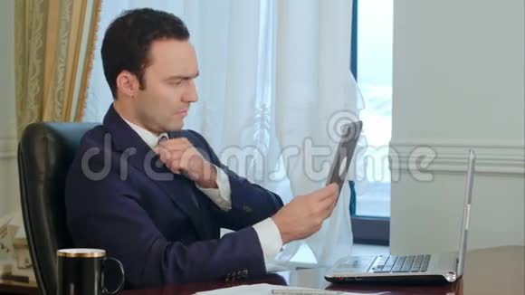一位优雅的商人正在整理他的西装外套和头发看着现代平板电脑视频的预览图