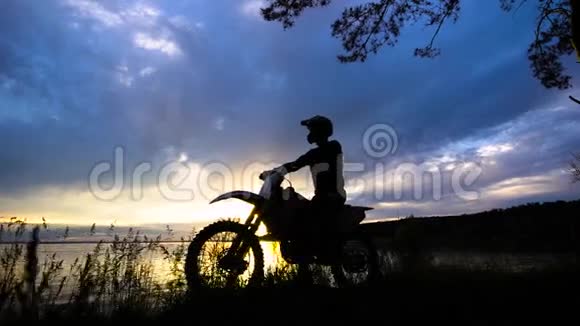 一位坐在摩托车上的极端交通司机的剪影一辆车站在夕阳背景下的河边视频的预览图