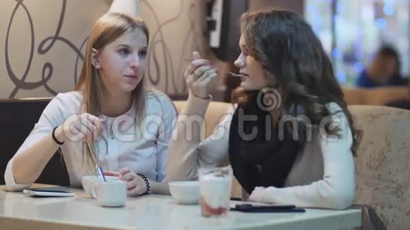 两个穿着白色衬衫的漂亮年轻女孩坐在咖啡馆的一张桌子旁一边吃一边说话视频的预览图