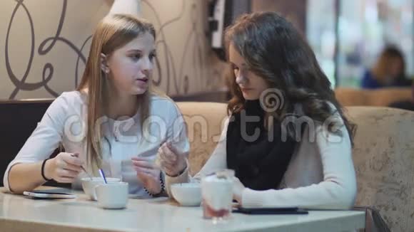 两个穿着白色衬衫的漂亮年轻女孩坐在咖啡馆的一张桌子旁一边吃一边说话视频的预览图