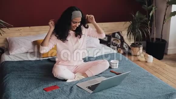 年轻迷人的女孩坐在床上玩笔记本电脑她唱歌跳舞高兴的好消息视频的预览图