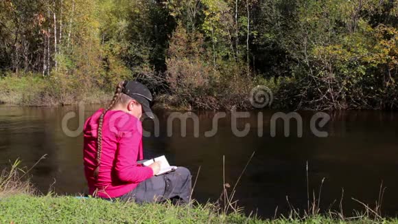 穿着粉红色连帽衫的女孩在河边的笔记本上做笔记有声音视频的预览图