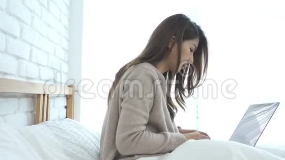 美丽的年轻微笑的亚洲女人在家里的卧室里坐在床上用笔记本电脑工作视频的预览图