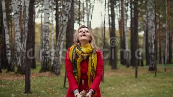 迷人的年轻女子抛出黄色的叶子脱落穿着风衣在秋天公园散步的女孩慢动作视频的预览图