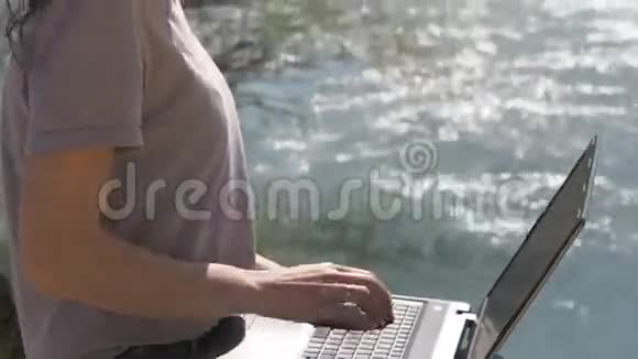 带着笔记本电脑的女孩在新鲜空气中工作一个女人正在河边的笔记本电脑上打字视频的预览图
