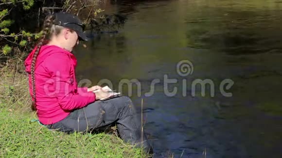 穿着粉红色连帽衫的女孩在河边的笔记本上做笔记有声音视频的预览图