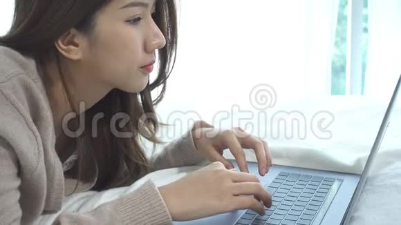 美丽的年轻微笑的亚洲女人在家里的卧室里坐在床上用笔记本电脑工作视频的预览图