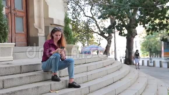 可爱的女孩看着她的手机坐在户外的台阶上视频的预览图
