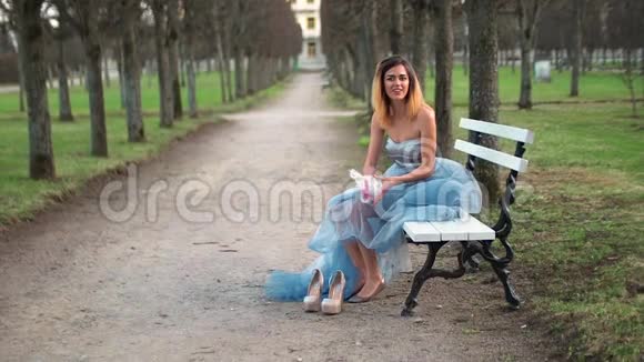 穿着银色和蓝色连衣裙的漂亮女孩坐在Parkway的长凳上调整着装准备在照片中摆姿势视频的预览图