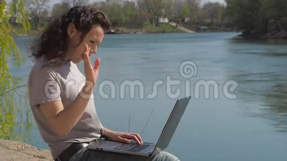 河边有台手提电脑的女孩一个年轻的女人在大自然中工作带笔记本电脑的水岸卷发女孩视频的预览图