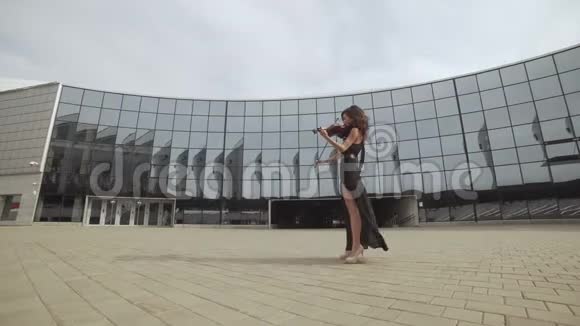 漂亮的女人穿着黑色连衣裙在玻璃大楼附近拉小提琴艺术概念视频的预览图