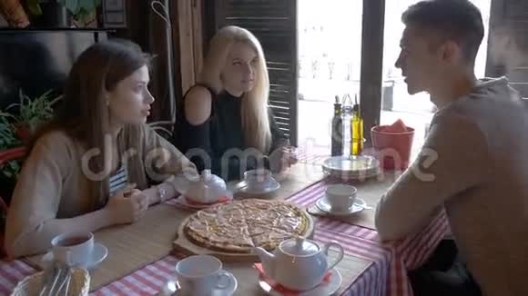 两个女孩和一个年轻人坐在一张桌子旁的咖啡馆里说话他们喝茶吃披萨他们告诉新闻他们视频的预览图