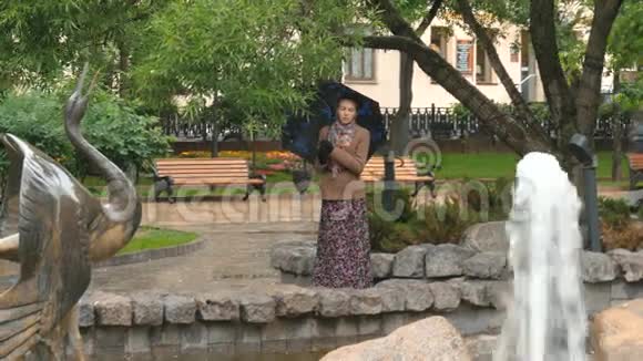 一个美丽的女孩悲伤的眼睛站在雨中的城市公园附近的喷泉视频的预览图