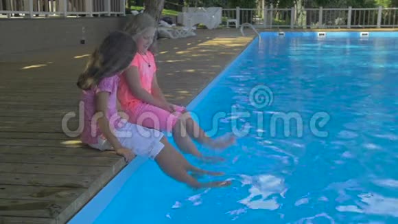 两个小女孩在游泳池里泡脚的户外镜头可爱的小女孩坐在游泳的边缘视频的预览图