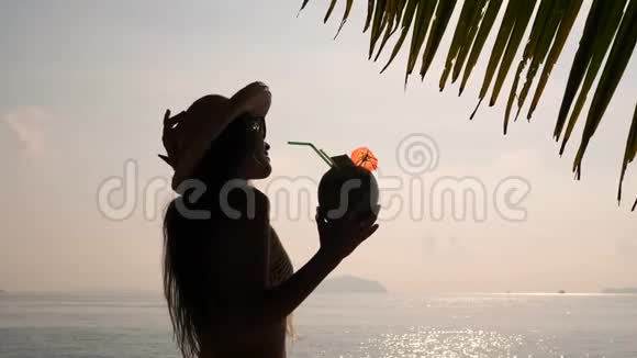 在热带海滩喝新鲜泰国椰子鸡尾酒的年轻旅游女孩日落剪影4K慢动作普吉岛视频的预览图