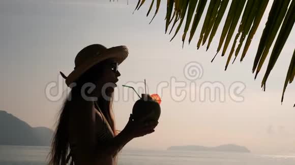 日落的剪影年轻的旅游女孩喝新鲜的泰国椰子水鸡尾酒在热带海滩4K慢行视频的预览图