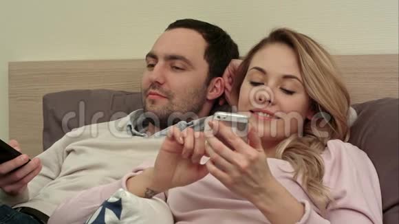 迷人的女人躺在床上对着手机发短信微笑着而她的伴侣则撬开屏幕视频的预览图