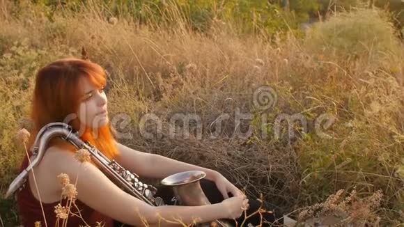 夕阳西下的剪影年轻漂亮的红发女孩在玩萨克斯管自然一个女人吹进烟斗概念爱好音乐视频的预览图