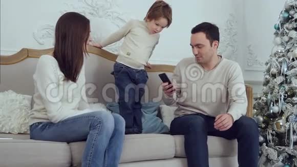 快乐的一家人和他们的儿子坐在沙发上而爸爸在打电话视频的预览图