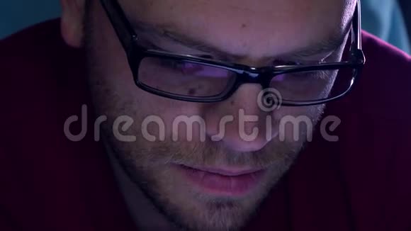 戴着黑边眼镜长着大胡子的年轻人在黑暗的房间里用他的平板电脑紫色的屏幕发光K4关闭视频的预览图