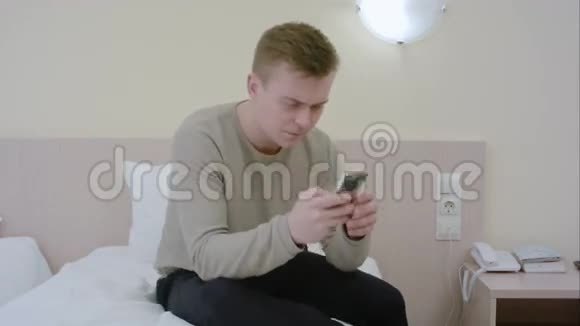 忧心忡忡的年轻人坐在家里的床上用手机打短信视频的预览图