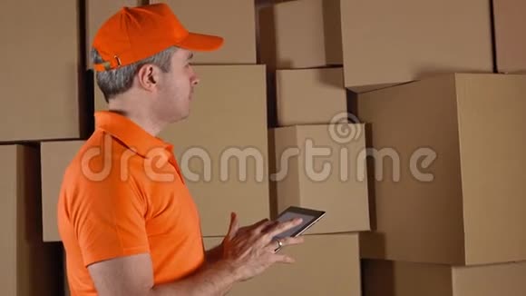 穿着橙色制服的仓库工人数着箱子用平板电脑对着棕色纸箱做记录4K演播室视频的预览图