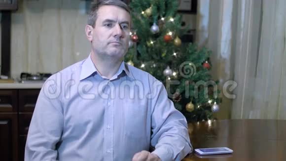 聪明的中年男人在圣诞树的背景下坐在家里的餐桌上用手机说话视频的预览图